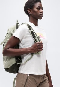 Άτομο που φοράει λευκό μπλουζάκι με τύπωμα και ανοιχτό καφέ παντελόνι, φορώντας ανοιχτοπράσινο σακίδιο στην μια ώμο, μπροστά σε απλό φόντο.