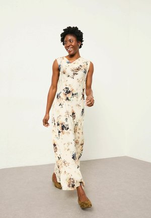 Floral-patterned, sleeveless jumpsuit in cream tones, featuring a draped neckline and flowing silhouette, paired with brown suede shoes.