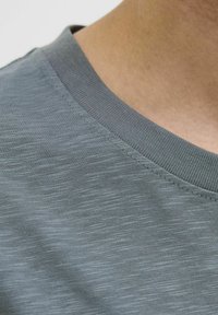 Camiseta de color gris claro con una textura suave, que presenta un cuello acanalado y un sutil patrón horizontal variado. Vista en primer plano de la tela.
