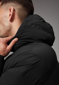 Chaqueta acolchada negra con capucha, que presenta una textura suave y un diseño ajustado. Primer plano del cuello y área del cuello, mostrando detalles de costura.