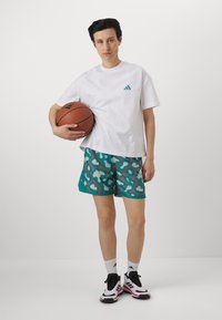 Biała bawełniana koszulka z zielonym logo, turkusowe szorty z abstrakcyjnym nadrukiem w panterkę, białe skarpetki oraz czarno-czerwone buty sportowe. Trzyma piłkę koszykową.