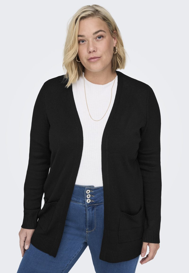 Zwarte cardigan met een open voorkant, geribbelde textuur en twee voorzakken, gedragen over een witte geribbelde top en blauwe high-waisted jeans.