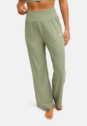 Vrouw die lichtgroene, geribbelde broek met wijde pijpen en een bijpassend cropped top draagt, staat blootsvoets tegen een effen achtergrond.