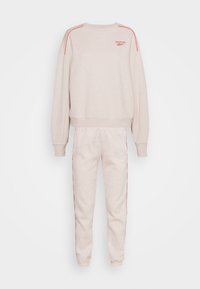 Survêtement beige clair Reebok avec sweat-shirt à manches longues et pantalon de jogging assorti avec un passepoil rouge et un logo sur la poitrine.