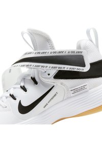 Baskets de sport blanches avec accents noirs, présentant un dessus texturé, une sangle "JUST DO IT" et une semelle en caoutchouc. Marque Nike visible.