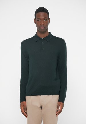 Tamnozeleni dukserica s dugim rukavima i pletenom teksturom, s tri gumba i klasičnim kragnom, u kombinaciji s bež hlačama.