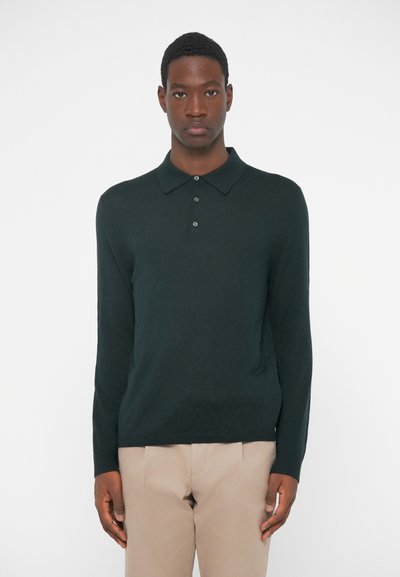 Camisa polo de manga larga verde oscuro con textura de punto, tapeta de tres botones y cuello clásico, combinada con pantalones beige.