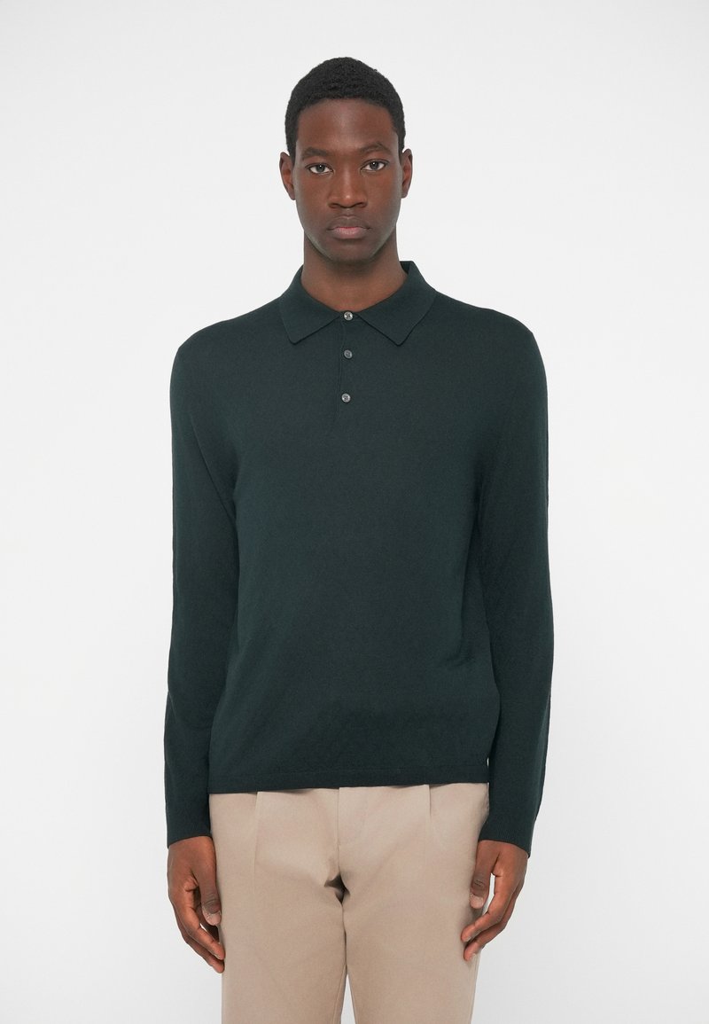 Σκούρο πράσινο πουκάμισο πόλο με μακριά μανίκια �και πλεκτή υφή, με τρεις κουμπάκια, και κλασικό γιακά, συνδυασμένο με μπεζ παντελόνι.