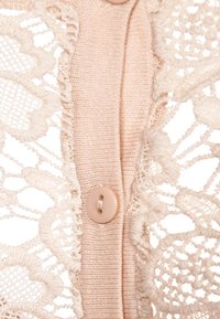 Gros plan sur un tissu maille beige avec un seul bouton beige et une bordure en dentelle florale délicate de chaque côté.