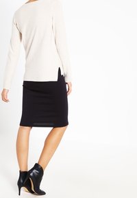 Frau, die einen cremefarbenen Pullover, einen schwarzen, knielangen Bleistiftrock und schwarze, hochhackige Stiefeletten trägt, steht mit leicht zur Seite gewendetem Rücken.