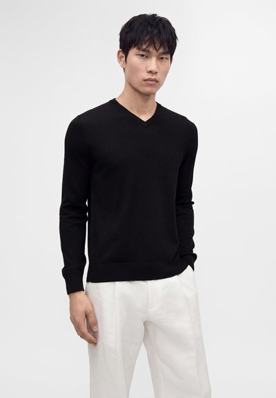 Homme portant un pull noir à manches longues en V et un pantalon blanc, debout sur un fond clair uni.