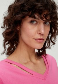 Różowa koszulka z dekoltem w serek z rozcięciami na szwach ramion, wykonana z miękkiego materiału, z luźnym krojem i subtelnymi detalami przeszycia.