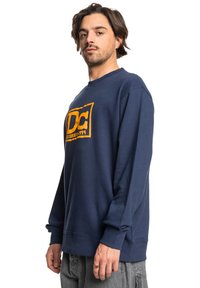Navy blue sweatshirt made from a soft fabric, featuring a bold orange DC logo on the front and ribbed cuffs and hem. Casual fit design.