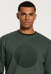 Groene sweatshirt van zacht materiaal, met een gestructureerd cirkelpatroon op de voorkant en een zwarte binnenkraag.