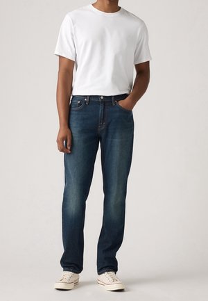 Hombre con camiseta blanca lisa de manga corta, vaqueros de mezclilla azul oscuro y zapatillas blancas, de pie con una mano en el bolsillo.