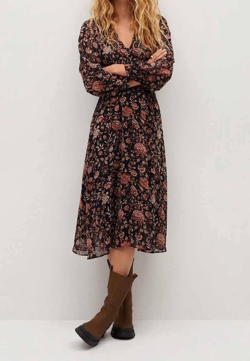 Femme aux longs cheveux blonds bouclés, vêtue d'une robe midi noire à fleurs et de bottes marron mi-mollet, debout les bras croisés contre un mur blanc.