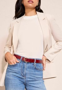 Beige blézer egy fehér, texturált felsőn, piros, fényes övvel, kerek arany csattal, kék farmernadrággal párosítva.