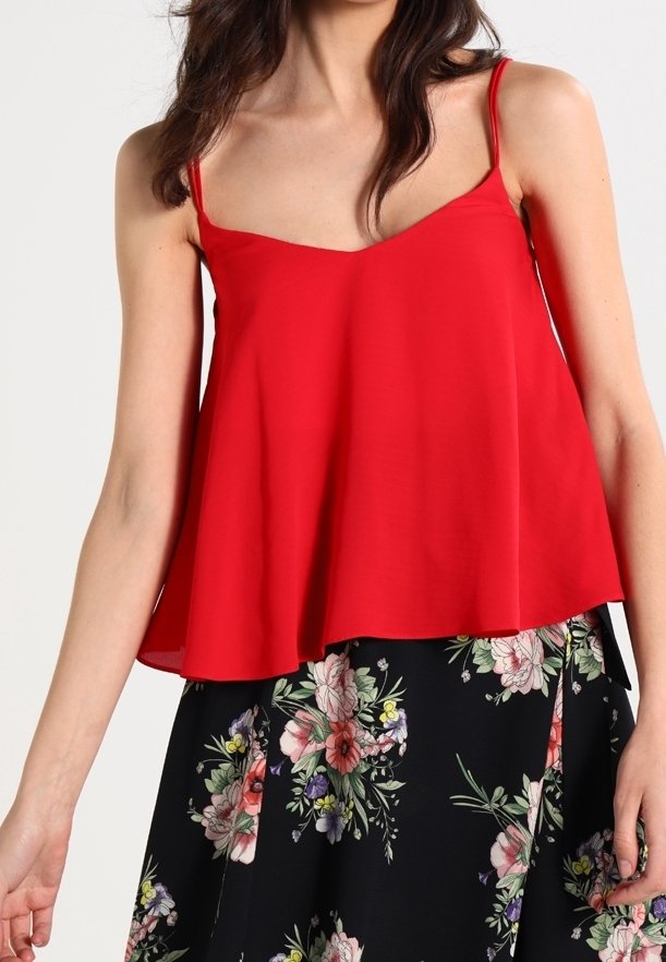 Femme portant un haut rouge sans manches à fines bretelles et une jupe noire avec un motif floral multicolore, debout sur fond blanc.