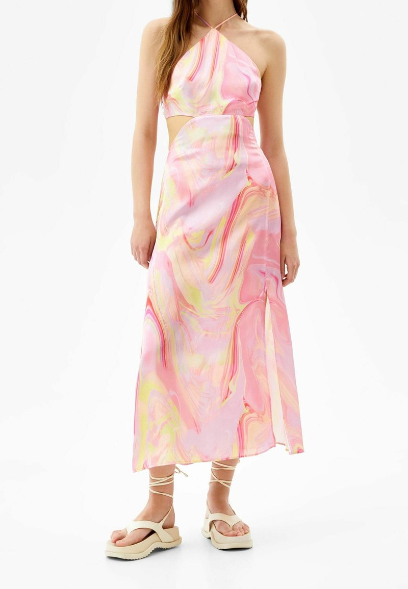 Femme portant une robe dos nu avec imprimé marbré rose et jaune, une découpe latérale et des sandales à plateforme beiges avec des brides à la cheville.