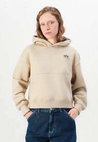 Felpa beige con tasca frontale, polsini a coste e un logo prominente sul petto. Indossata con pantaloni di jeans blu scuro.