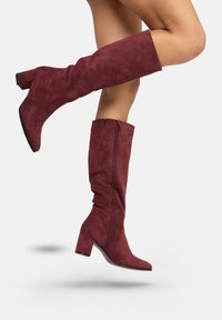 Botas de ante burdeos hasta la rodilla con un tacón bloque moderado, punta afilada y detalle de cremallera lateral para un fácil uso. Textura suave.