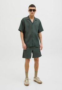 Chemise à manches courtes vert foncé avec des rayures verticales, assortie à un short. Portée avec des baskets beiges et des chaussettes de longueur chevillère. Lunettes de soleil carrées.