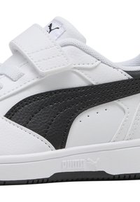 Baskets blanches avec des accents en cuir noir, sangle Velcro, dessus texturé et semelle en caoutchouc. Présente des détails brodés et un logo sur le côté.