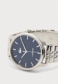 Ασημένιο μεταλλικό ρολόι χειρός με βαθύ μπλε καντράν, λευκούς δείκτες ώρας, τρεις δείκτες, το λογότυπο της Lacoste και μπρασελέ από συνδεδεμένα μεταλλικά κρίκους πάνω σε λευκό φόντο.