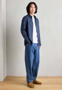 Camisa de botones azul marino con un patrón de cuadros azules, sobre una camiseta blanca. Combinada con pantalones vaqueros anchos azules y zapatos estilo mocasín marrones.