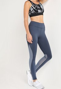 Collant da donna blu navy a vita alta con una superficie liscia. Presentano pannelli laterali in un grigio chiaro a contrasto e una vestibilità aderente. Abbinati a un reggiseno sportivo bianco e nero.
