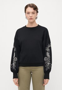 Kvinne iført en svart hettegenser med hvite broderte blomsterdesign på ermene og olivengrønne bukser, som står mot en ensfarget bakgrunn.