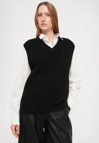 Mustan ribbed neule sleeveless-pullovers V-kauluksella, valkoisen kauluspaidan päällä, yhdistetty mustiin keinonahkahousuihin.