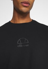 Primer plano de una persona que lleva una camiseta negra de cuello redondo con un sutil logo de ellesse en relieve en el pecho.
