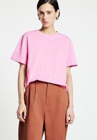 T-shirt di cotone rosa con maniche corte, scollo rotondo e vestibilità comoda, abbinata a pantaloni marroni a vita alta e anelli d'argento.