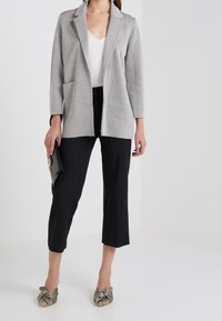 Femme portant un blazer argenté pailleté, un top blanc, un pantalon court noir, des mules pointues métalliques et tenant un sac à main noir.