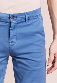 Světle modré bavlněné kalhoty s knoflíkovým zapínáním, poutky na opasek a hladkým předním dílem. Obsahují jemné prošívání a hladkou texturu.