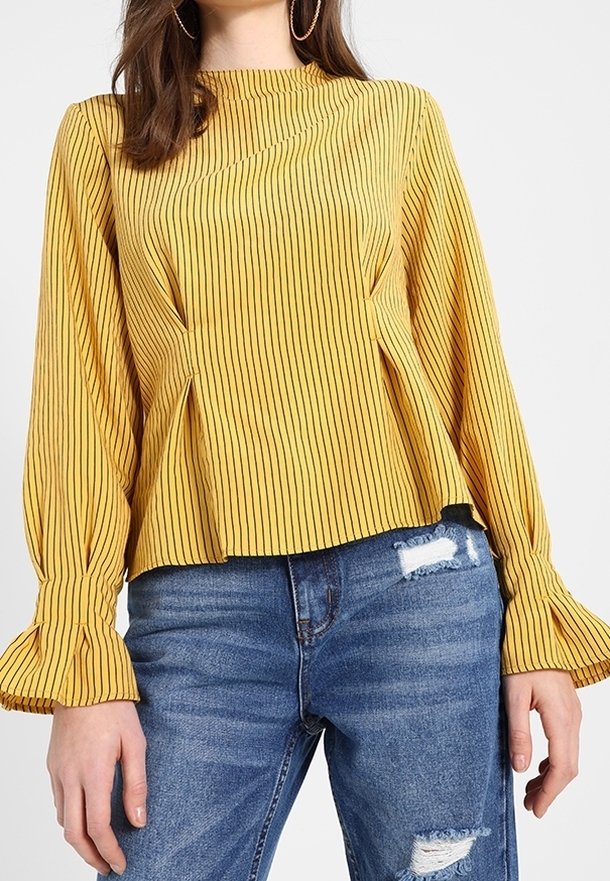 Blouse rayée jaune avec manches longues en cloche et détails plissés, associée à un jean bleu déchiré. Le haut a un col montant et une coupe décontractée.