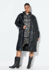 Cappotto in faux leather nero con chiusura a zip frontale, maniche lunghe e polsini elasticizzati, indossato sopra un vestito a camicia con stampa leopardata e stivali da cowboy neri.