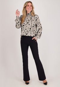 Lichtgrijze blouse met zwarte polkadots en een geplooide kraag, gecombineerd met zwarte broek met gouden knoopaccenten. Zwarte hakken maken de outfit compleet.