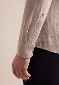 Camicia in lino a maniche corte di colore beige chiaro con una tessitura a rilievo, caratterizzata da bottoni bianchi e un dettaglio ricamato a forma di fiore sul polsino.