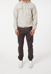 Ljusgrå zip-up jacka med huva, parat med mörka cargobyxor med sidofickor och elastiska muddar. Vita sneakers fulländar outfiten.