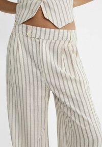 Pruhované kalhoty v krémové a šedé barvě, vyrobené z lehké tkaniny. Mají plisování, zapínání na knoflík a boční kapsy pro přidané detaily.