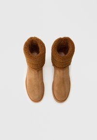 Botine de iarnă din suede maro, cu căptușeală groasă din fleece pliată în jurul vârfului, văzute de sus pe un fundal alb.