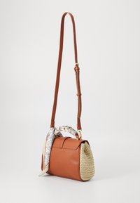 Brązowa skórzana torba na ramię z plecionego siana z regulowanym paskiem oraz wiązaną białą, kwiatową chustą przy uchwycie, na białym tle.