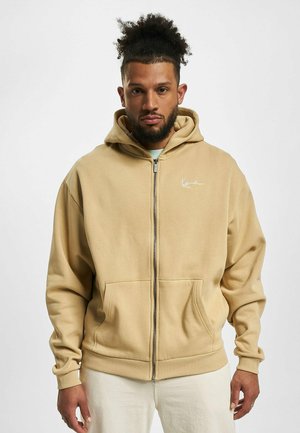 Beige zip-up hette laget av mykt stoff, med en frontlomme, ribbestrikkede mansjetter og en minimalistisk logo på brystet.