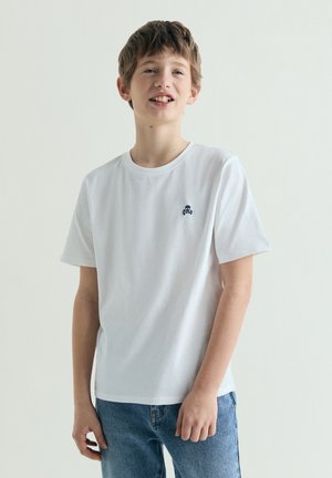 Junger Junge mit kurzen braunen Haaren, der ein weißes T-Shirt mit kleinem Totenkopf-Logo und blaue Jeans trägt, steht vor einem schlichten hellen Hintergrund.