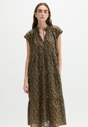 Jeune femme aux longs cheveux bruns porte une robe sans manches, ample, vert foncé avec un petit imprimé floral et un décolleté froncé.