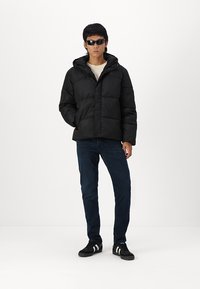 Black puffer jacket with a hood, featuring quilted design and front closure. Paired with dark jeans and black shoes with white stripes.