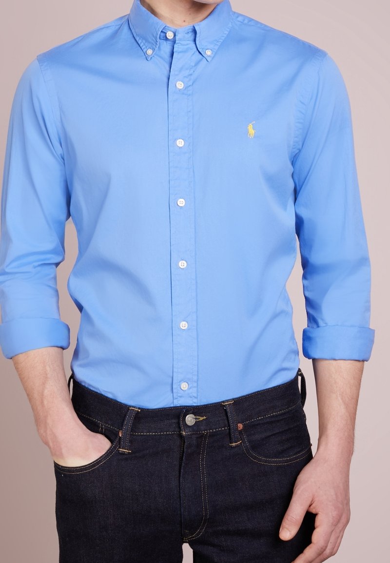 Světlomodrá košile na knoflíky s límcem, dlouhými rukávy vyhrnutými na manžetách a malým žlutým logem na hrudi. Nosená s tmavými džíny.