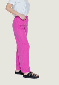 Pantaloni in lino rosa con vita elastica e cordoncino. Abbinati a sandali neri con fibbia. Il tessuto appare leggero e casual.